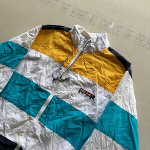 May include: A color-blocked windbreaker jacket featuring horizontal panels of white, mustard yellow, and teal, with black accents at the shoulders. The jacket has a high collar, a full-length front zipper, and a small embroidered nautical flag logo.