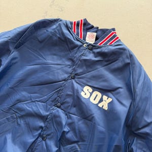 May include: A blue nylon snap-front jacket featuring the white text SOX on the chest. The garment has a ribbed collar with red and white stripes, long sleeves, and dark blue snap buttons. A white interior tag is visible at the neckline.