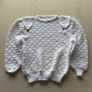Puede incluir: Un suéter blanco de manga larga con un patrón de tejido festoneado texturizado en todo el cuerpo y las mangas. El suéter incluye dos solapas en los hombros, cada una asegurada con un pequeño botón circular multicolor en el borde.