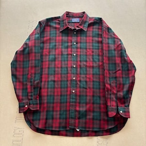 Puede incluir: Una camisa de manga larga con botones y un patrón de cuadros en rojo y verde oscuro. La camisa incluye un cuello puntiagudo, un bolsillo en el pecho en el lado izquierdo y puños abotonados. La tapeta frontal muestra una serie de botones.