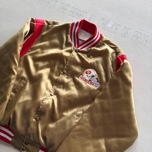 May include: A gold satin bomber jacket featuring red accents on the sleeves and collar. The jacket has a snap-button front closure and a San Francisco 49ers football helmet logo patch on the left chest with the text 49ERS and an NFL logo.