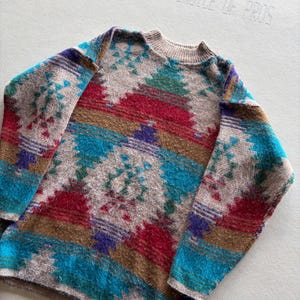 May include: A long-sleeve knit sweater featuring a textured, multi-colored geometric pattern in shades of tan, bright red, teal, purple, and brown. The garment has a ribbed mock neck collar and a fuzzy, soft-touch finish across the entire surface.