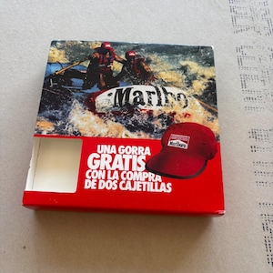 May include: A rectangular cardboard promotional box featuring a graphic of two people whitewater rafting. The box displays a red baseball cap with a Marlboro logo and the Spanish text: UNA GORRA GRATIS CON LA COMPRA DE DOS CAJETILLAS.