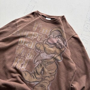 Op de afbeelding: Een bruin sweatshirt met ronde hals en een grafische print van het personage Grumpy uit Sneeuwwitje. De tekst "I'm Grumpy! Deal With It!" is in een verweerd wit lettertype gedrukt naast het personage met gekruiste armen en een frons.