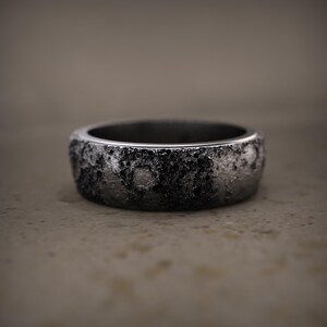Puede incluir: Un anillo plateado texturizado con una superficie oscura y moteada. El anillo tiene una banda ancha y un interior liso. El diseño le da al anillo una estética robusta e industrial. El anillo es una joya.