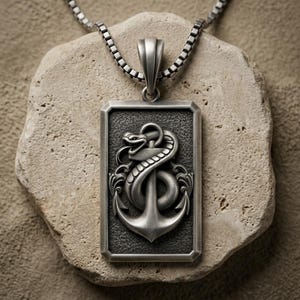 May include: A rectangular silver-toned pendant featuring a raised relief of a coiled snake wrapped around a ship anchor. The pendant has a textured dark gray background and is suspended from a silver-toned box chain necklace.