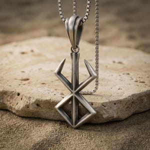 Puede incluir: Un colgante de metal plateado con el símbolo de la Marca del Sacrificio, suspendido de una cadena de eslabones cuadrados a juego. El colgante tiene un acabado mate cepillado con líneas angulares nítidas y una anilla prominente en la parte superior.