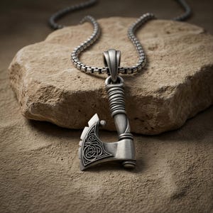 Puede incluir: Un colgante de metal plateado con forma de hacha de batalla, que presenta intrincados grabados de nudos celtas en la hoja y un diseño de mango envuelto. El colgante cuelga de una cadena de eslabones cuadrados a juego con acabado pulido.