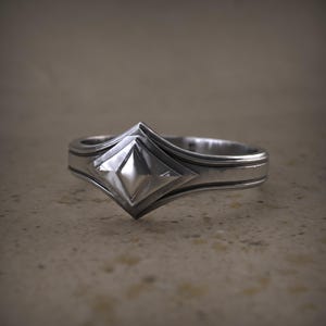 Peut inclure: Une bague en argent avec un motif central en forme de losange géométrique. La bague présente une bande texturée avec un design subtil de rainures. La forme de diamant est en relief et reflète la lumière.