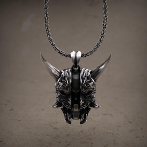 May include: A silver-colored pendant necklace featuring a detailed oni mask design. The mask has large horns, intricate facial features, and is suspended from a matching chain. The pendant is a striking piece of jewelry.