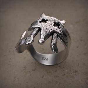 May include: A silver-colored ring featuring a detailed wolf head design. The ring has an open design, with the wolf's head at the top and the band curving around. The ring is marked with "925".