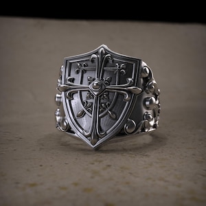 Könnte beinhalten: Ein silberner Ring mit einem schildförmigen Design, verziert mit einem Kreuz und floralen Details. Der Ring hat ein aufwendiges Muster und ein breites Band. Die Schildform ist zentral, mit einem Kreuz und dekorativen Elementen.