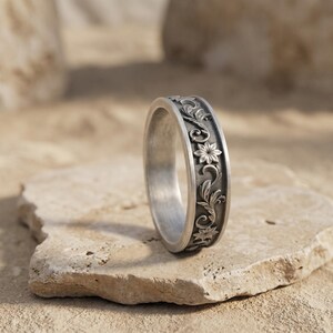 May include: A silver-toned metal ring featuring an intricate, raised floral and vine relief pattern along its outer band. The surface of the ring has a matte, brushed finish with darkened recessed areas that highlight the detailed botanical carvings.