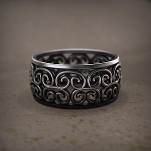 May include: A silver ring featuring an elaborate openwork design of swirling motifs. The ring has a broad band with a polished upper edge and a dark base, creating a contrast. The ring is likely made of metal and is suitable for everyday wear.