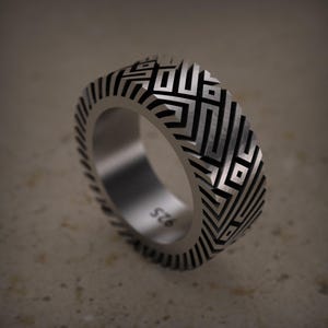 May include: A silver ring with a geometric pattern. The ring features a repeating design of black lines and shapes, creating a textured appearance. The band is approximately 0.5 inches wide and has the number "925" engraved on the inside.