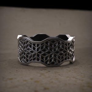 May include: A silver ring featuring a detailed floral pattern. The ring has a wide band with a repeating geometric design. The edges of the ring have a wavy design. The ring is displayed on a neutral surface.