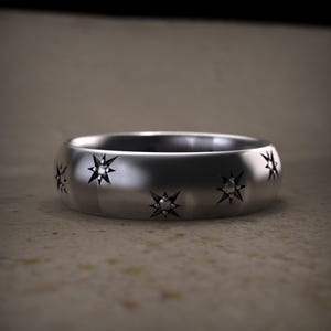 May include: A silver ring with a polished finish. The ring features engraved starburst designs around the band. The ring is a simple band style, with a smooth, rounded surface. The ring is likely made of a metal such as sterling silver or stainless steel.