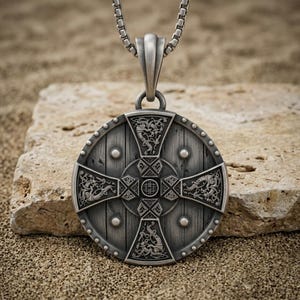May include: A circular silver-toned pendant featuring a detailed shield design with a central cross, intricate knotwork, and engraved animal motifs. The pendant is suspended from a silver-toned box chain, showcasing a weathered metallic finish.