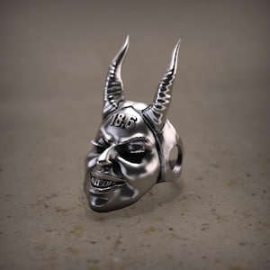 May include: A silver ring featuring a devil face design with curved horns and the number "18.6" engraved on the forehead. The ring has a detailed, expressive face with a sinister grin.