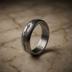 May include: A silver ring with engraved script, possibly Elvish, encircles the band. The ring has a brushed metal finish and the number "925" is visible. The ring is set against a neutral, marble-like surface.