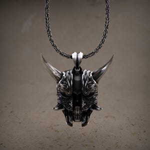 May include: A silver-colored pendant necklace featuring a detailed oni mask design. The mask has large horns, intricate facial features, and is suspended from a matching chain. The pendant is a striking piece of jewelry.