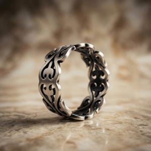 Könnte beinhalten: Ein silberner Ring mit einem aufwendigen, durchbrochenen Design. Der Ring weist ein sich wiederholendes Muster aus stilisierten Blatt- oder Flammenformen auf, das ein durchgehendes Band bildet. Das Metall hat ein leicht oxidiertes Aussehen, das die Details des Designs hervorhebt.