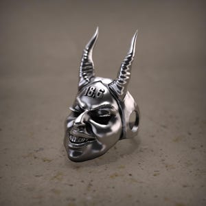 Peut inclure: Une bague argentée représentant un visage de diable avec des cornes incurvées et le chiffre "18.6" gravé sur le front. La bague présente une surface détaillée et texturée et une expression sinistre.