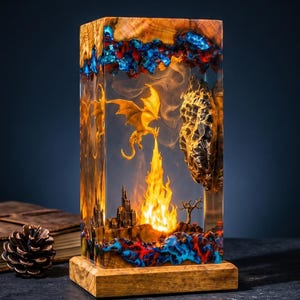 Puede incluir: Una lámpara rectangular de madera y resina transparente con una figura de dragón dorado que exhala fuego sobre un pequeño castillo. Los bordes superior e inferior tienen detalles en resina azul y roja, y el interior emite una luz cálida.