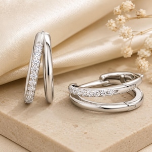 May include: Silver hoop earrings, one with a row of small, clear gemstones. The other is a double hoop design, also with gemstones. The earrings are displayed on a neutral surface, with a soft, cream-colored background.