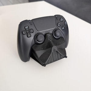 Könnte beinhalten: Ein schwarzer Videospiel-Controller auf einem schwarzen, 3D-gedruckten Ständer in Form einer Helmmaske. Der Controller verfügt über ein zentrales Touchpad, zwei Analogsticks, ein Steuerkreuz und vier Aktionstasten mit Dreieck-, Kreis-, Kreuz- und Quadrat-Symbolen.
