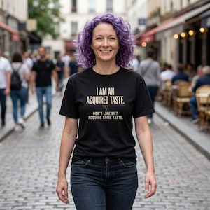 Pode incluir: Camiseta preta com o texto "I AM AN ACQUIRED TASTE." e a frase "DON'T LIKE ME? ACQUIRE SOME TASTE." impressa em branco. A pessoa usa jeans azuis e tem cabelo roxo.
