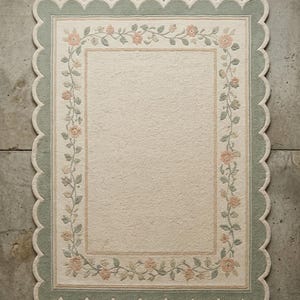 May include: A rectangular area rug featuring a cream-coloured centre bordered by a floral vine pattern with peach flowers and sage green leaves. The rug has a wide sage green scalloped edge and a cream-coloured trim, creating a decorative border.