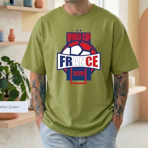 May include: A heather green short-sleeve t-shirt featuring a graphic of a soccer ball and the text "WORLD CUP FRANCE 2026" in red, white, and blue. The design is centered on the chest, showcasing a bold, athletic style with a round neckline.