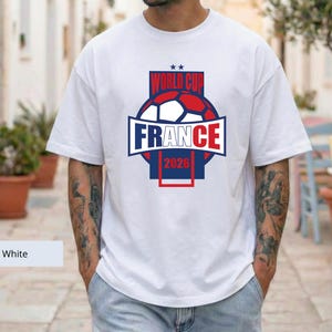 May include: A white short-sleeved t-shirt featuring a graphic design on the chest. The design includes a soccer ball in red, white, and blue, with the text "WORLD CUP" above it and "FRANCE 2026" below, all enclosed within a rectangular frame.