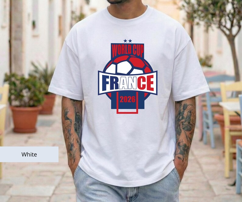 May include: A white short-sleeved t-shirt featuring a graphic design on the chest. The design includes a soccer ball in red, white, and blue, with the text "WORLD CUP" above it and "FRANCE 2026" below, all enclosed within a rectangular frame.