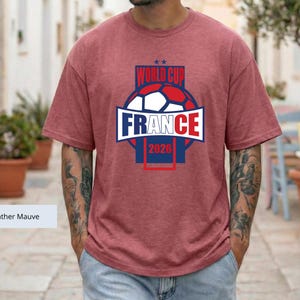 May include: A heather mauve short-sleeve t-shirt featuring a graphic of a soccer ball and the text "WORLD CUP FRANCE 2026" in blue, white, and red. The design includes two small stars above the text and a rectangular frame surrounding the graphic.