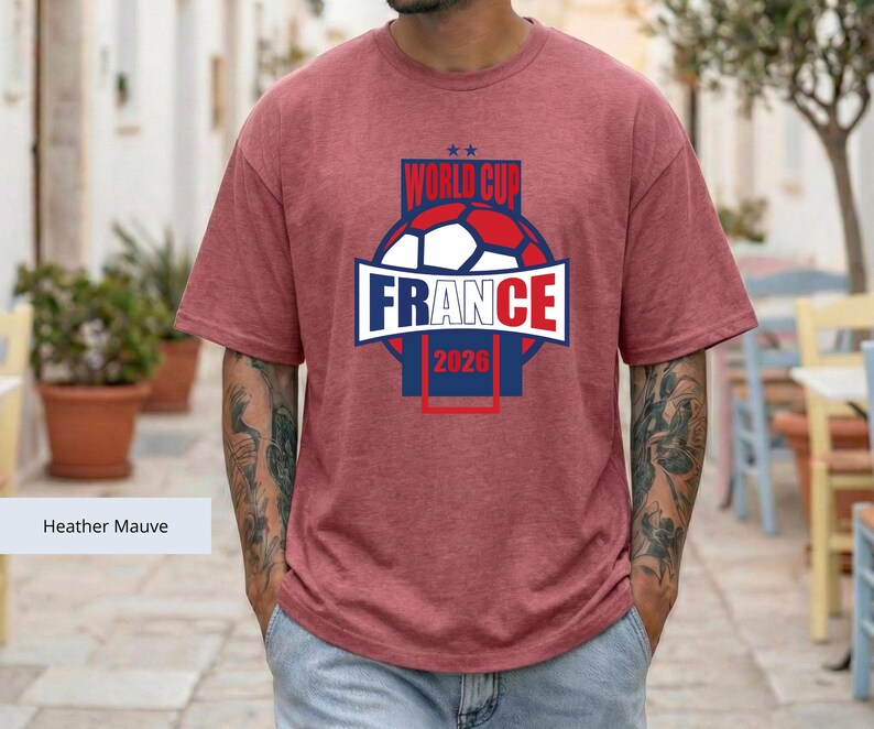 May include: A heather mauve short-sleeve t-shirt featuring a graphic of a soccer ball and the text "WORLD CUP FRANCE 2026" in blue, white, and red. The design includes two small stars above the text and a rectangular frame surrounding the graphic.