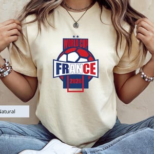 May include: A natural-colored short-sleeve t-shirt featuring a graphic print of a soccer ball in red, white, and blue. The text reads WORLD CUP FRANCE 2026, with two small stars above the text. The design is centered on the chest of the shirt.