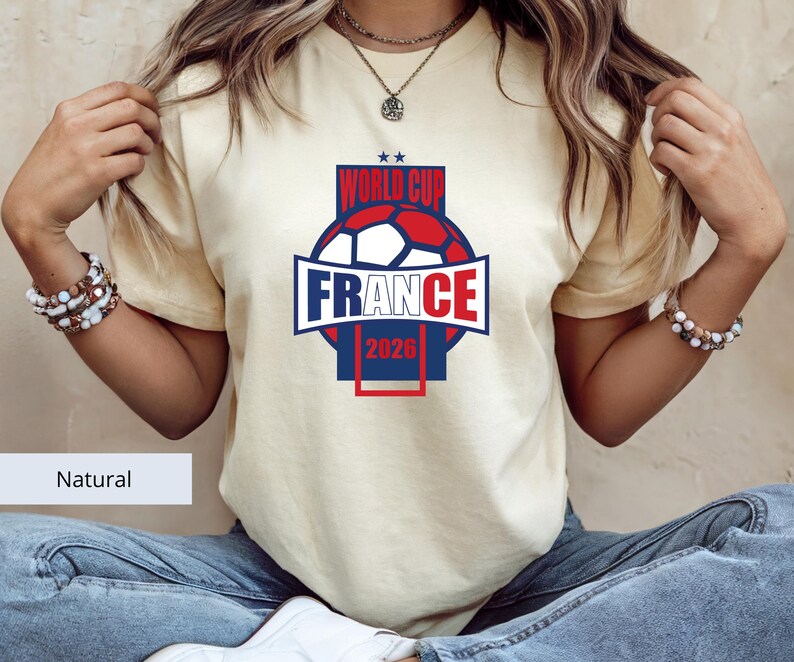 May include: A natural-colored short-sleeve t-shirt featuring a graphic print of a soccer ball in red, white, and blue. The text reads WORLD CUP FRANCE 2026, with two small stars above the text. The design is centered on the chest of the shirt.