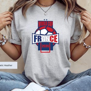 May include: An athletic heather gray short-sleeve t-shirt featuring a graphic design with a soccer ball, the text "WORLD CUP", "FRANCE", and "2026" in blue and red, with two small blue stars above the text, centered on the chest area.