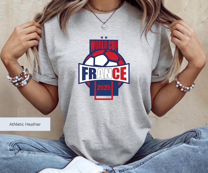 May include: An athletic heather gray short-sleeve t-shirt featuring a graphic design with a soccer ball, the text "WORLD CUP", "FRANCE", and "2026" in blue and red, with two small blue stars above the text, centered on the chest area.