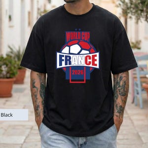 May include: A black short-sleeve t-shirt featuring a graphic design with a soccer ball and the text "WORLD CUP FRANCE 2026" in red, white, and blue colors. The design is centered on the chest and includes two small stars above the text.