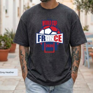 May include: A dark grey heather short-sleeve t-shirt featuring a graphic of a soccer ball with the text "WORLD CUP" above it and "FRANCE 2026" below it. The design uses red, white, and blue colors, with the word "FRANCE" centered in a white banner.