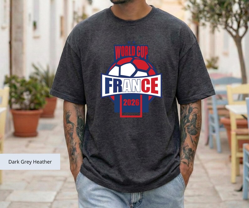 May include: A dark grey heather short-sleeve t-shirt featuring a graphic of a soccer ball with the text "WORLD CUP" above it and "FRANCE 2026" below it. The design uses red, white, and blue colors, with the word "FRANCE" centered in a white banner.