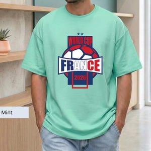 May include: A mint green short-sleeved t-shirt featuring a graphic on the chest. The design includes a soccer ball, the text "WORLD CUP", "FRANCE", and "2026" in red, white, and blue colors, with two small stars positioned above the text.