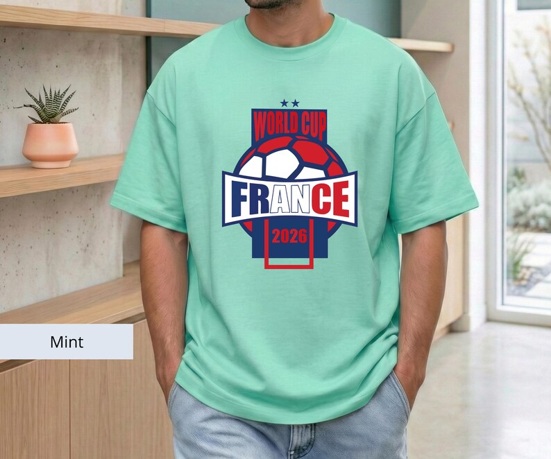 May include: A mint green short-sleeved t-shirt featuring a graphic on the chest. The design includes a soccer ball, the text "WORLD CUP", "FRANCE", and "2026" in red, white, and blue colors, with two small stars positioned above the text.