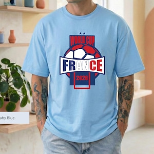 May include: A baby blue short-sleeve t-shirt featuring a graphic design with a soccer ball, the text "WORLD CUP", "FRANCE", and "2026" in red, white, and blue, with two small stars above the text, centered on the chest of the garment.