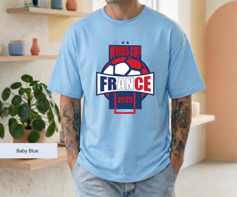 May include: A baby blue short-sleeve t-shirt featuring a graphic design with a soccer ball, the text "WORLD CUP", "FRANCE", and "2026" in red, white, and blue, with two small stars above the text, centered on the chest of the garment.