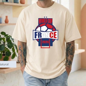 May include: A natural-colored short-sleeve t-shirt featuring a graphic design on the chest. The design includes a soccer ball, the text "WORLD CUP", "FRANCE", and "2026" in blue and red colors, with two small stars above the text.