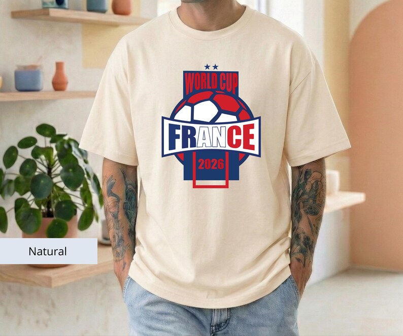 May include: A natural-colored short-sleeve t-shirt featuring a graphic design on the chest. The design includes a soccer ball, the text "WORLD CUP", "FRANCE", and "2026" in blue and red colors, with two small stars above the text.